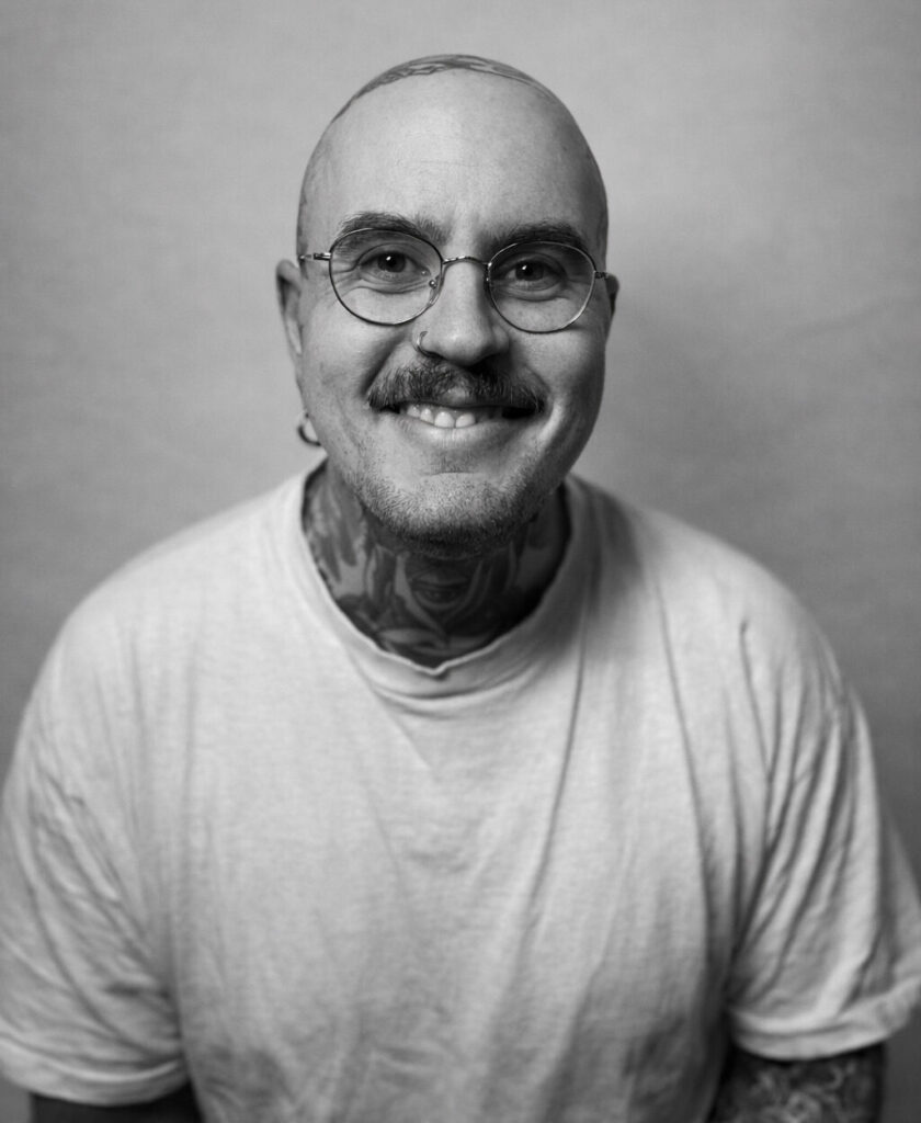 Ein lächelnder Mann mit Glatze, Brille und auffälligen Tattoos trägt ein weißes Shirt