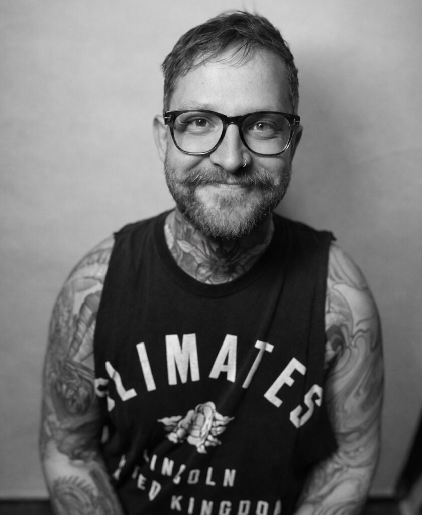 Ein lächelnder Mann mit Brille, Bart und vielen Tattoos trägt ein dunkles Shirt